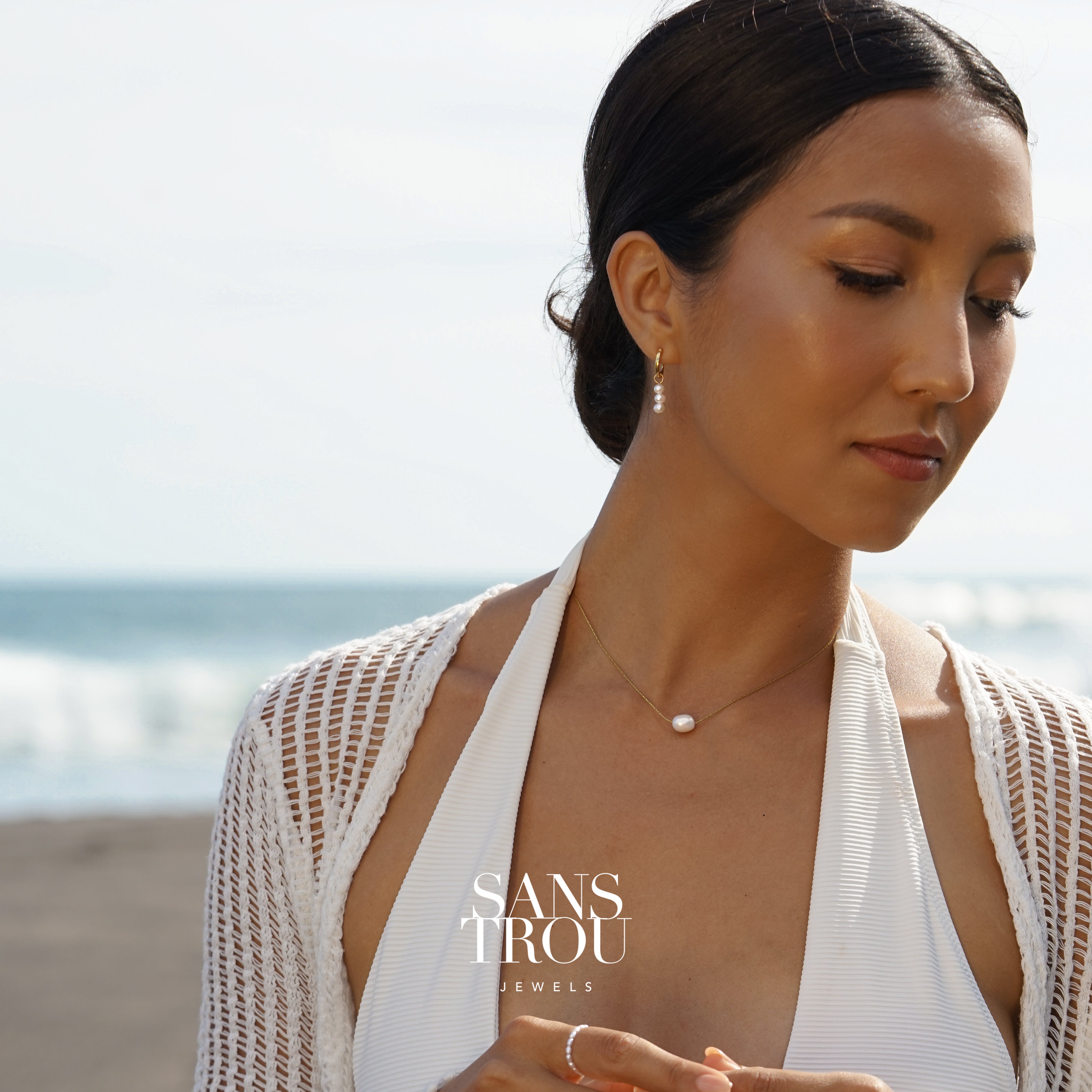 Woman wearing a dainty floating pearl necklace on a shiny gold chain, blurred beach in background