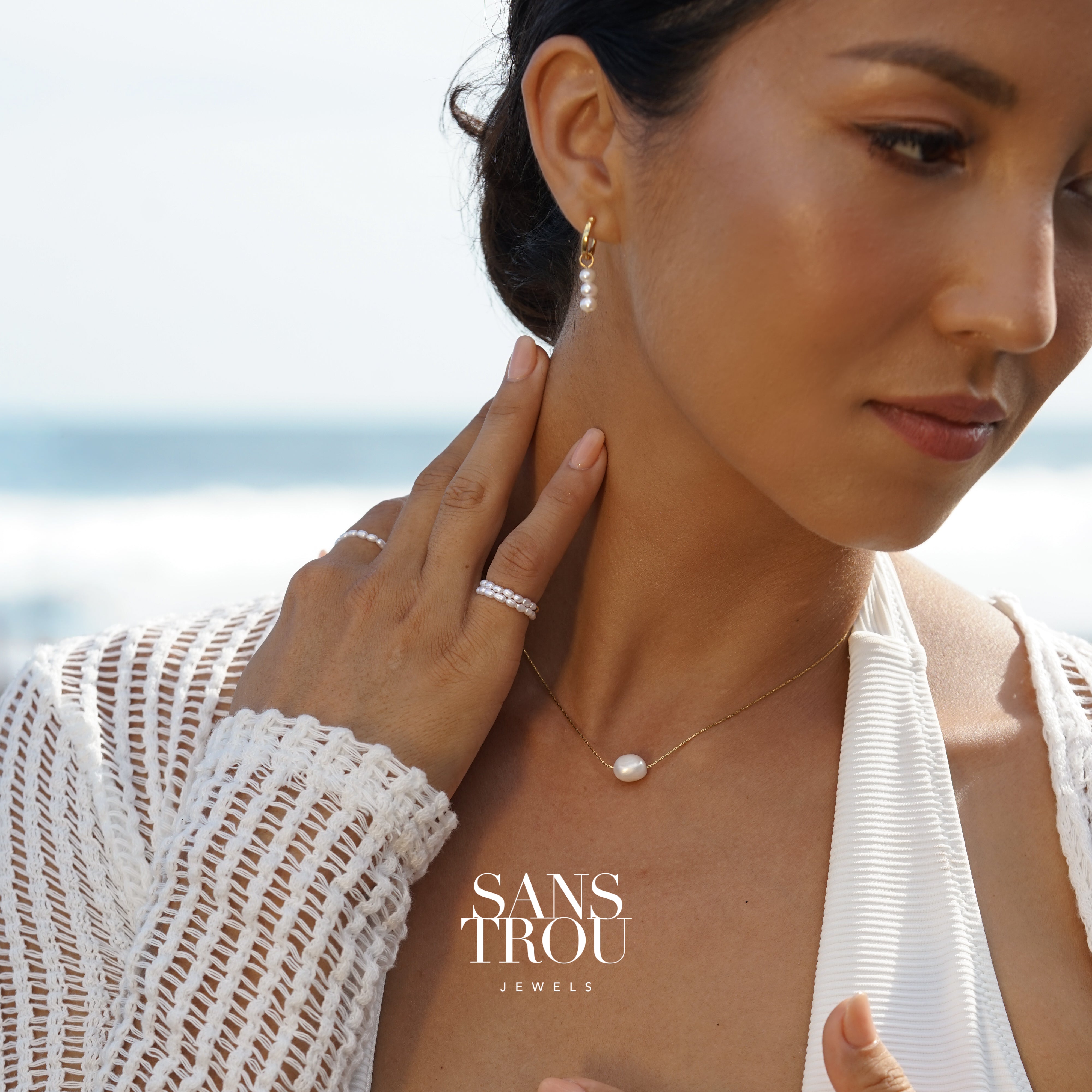 Woman wearing a dainty floating pearl necklace on a shiny gold chain, blurred beach in background