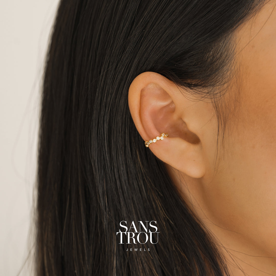 Model wearing a 18k gold plated ear cuff with CZ stones arranged in an asymmetrical composition.  Model wears the cuff on the conch. 