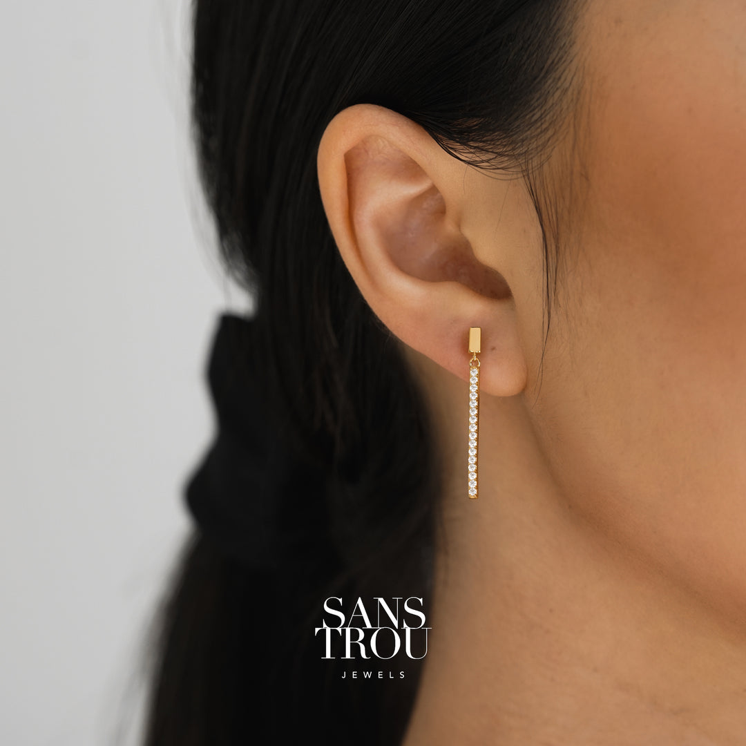 Model wears 18k gold plated clip-on drop earrings featuring a slim sterling silver bar with CZ stones. 